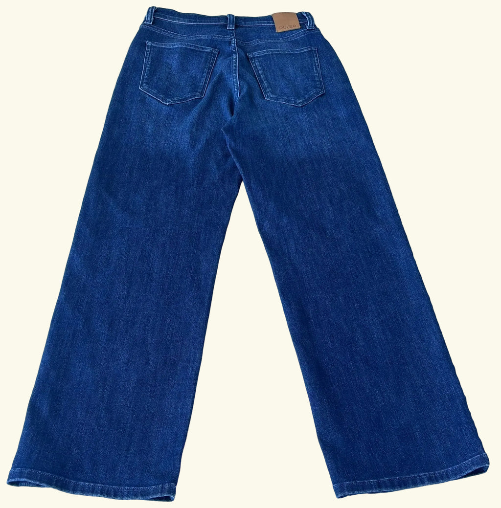 A pair of dark blue denim jeans shown from the back, laid flat on a light-colored tiled floor. The jeans feature a classic five-pocket design and a small tan leather brand patch at the back waistband.