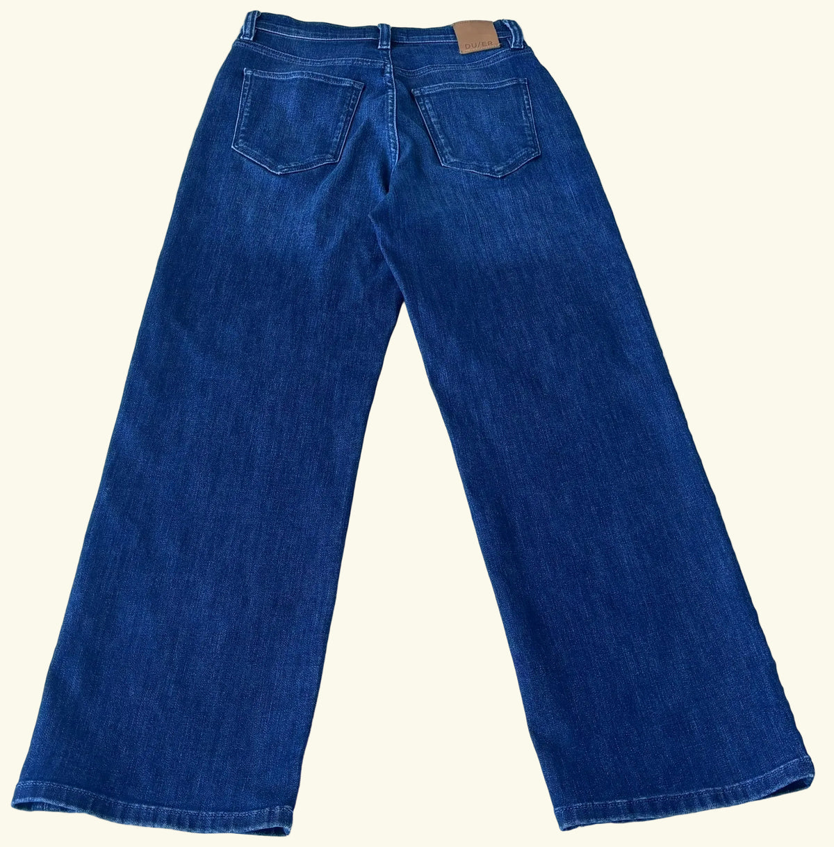 A pair of dark blue denim jeans shown from the back, laid flat on a light-colored tiled floor. The jeans feature a classic five-pocket design and a small tan leather brand patch at the back waistband.
